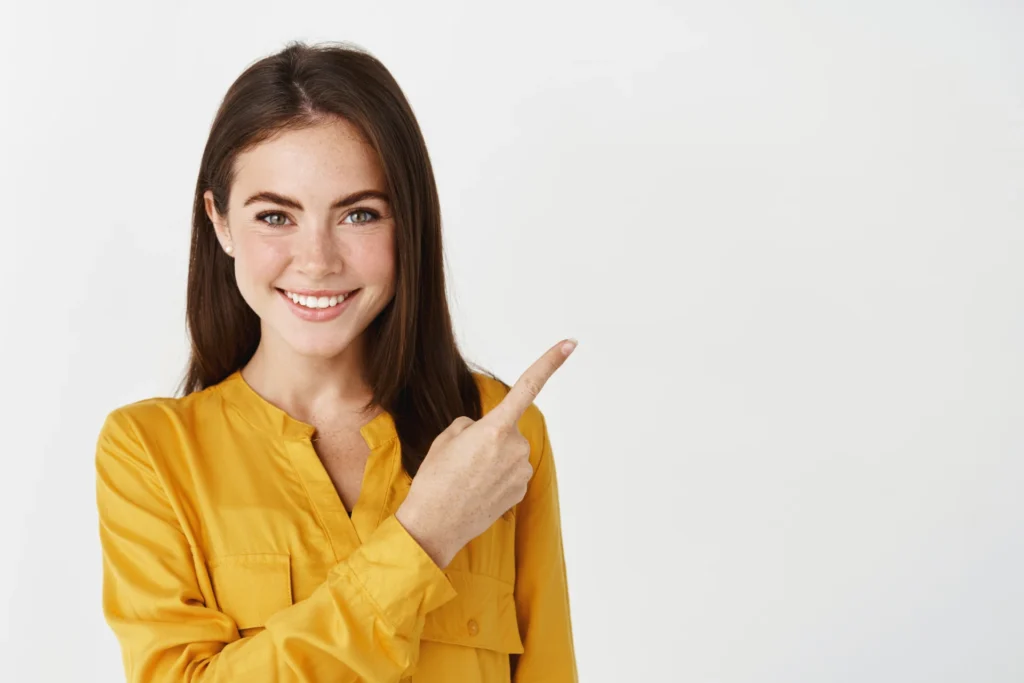 A woman with long, dark brown hair and a yellow blouse is smiling and pointing to her right